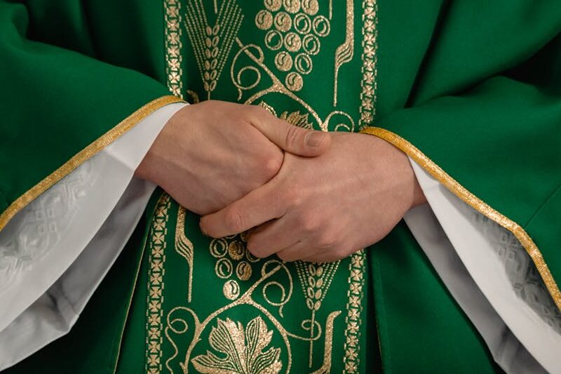 A close-up view of a priest's hands clasped in prayer wearing a green chasuble with gold embroidery.